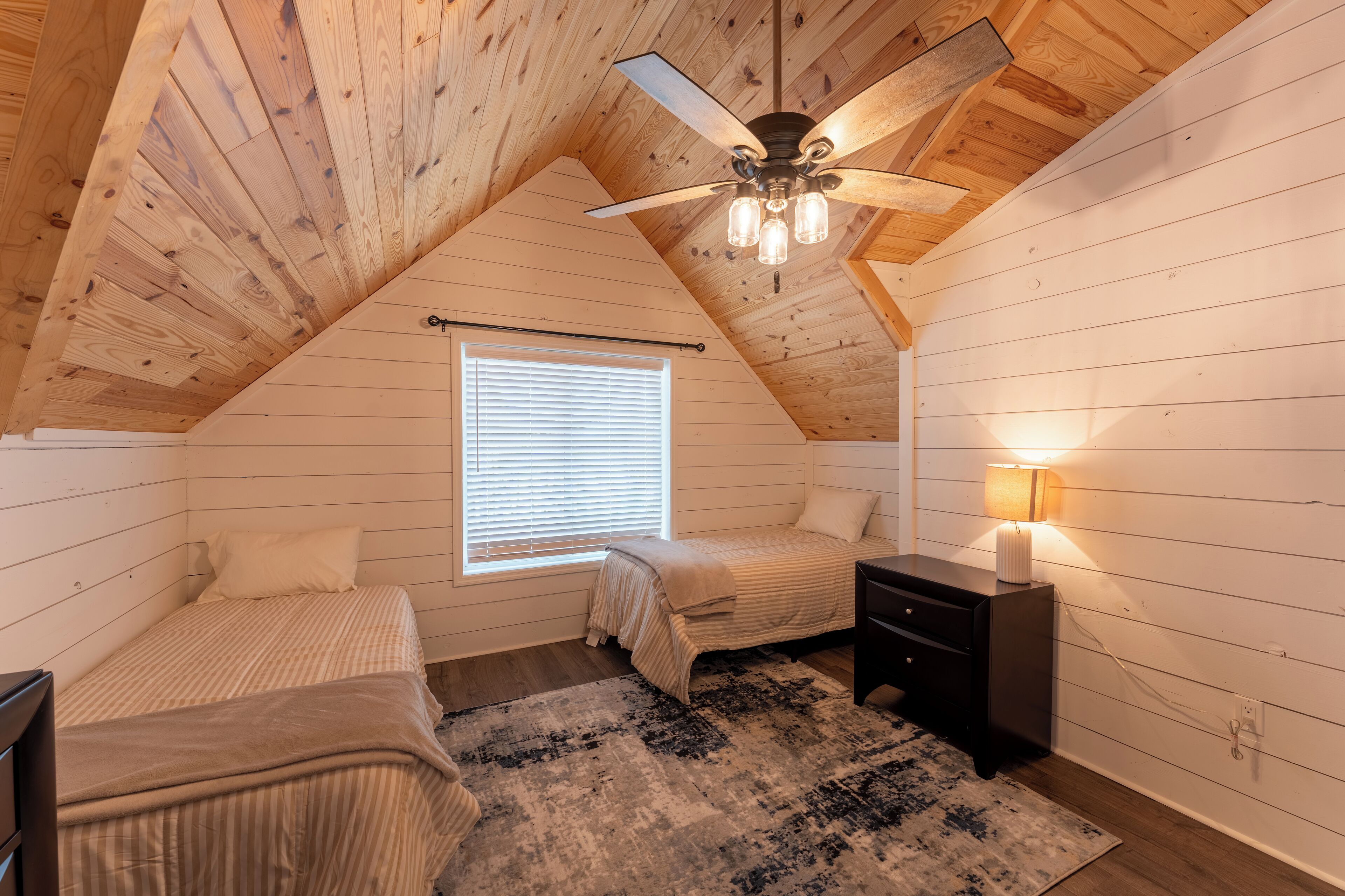 A cozy attic bedroom with wooden walls, a sloped ceiling, a ceiling fan, a window with blinds, two beds, a nightstand, and a rug.