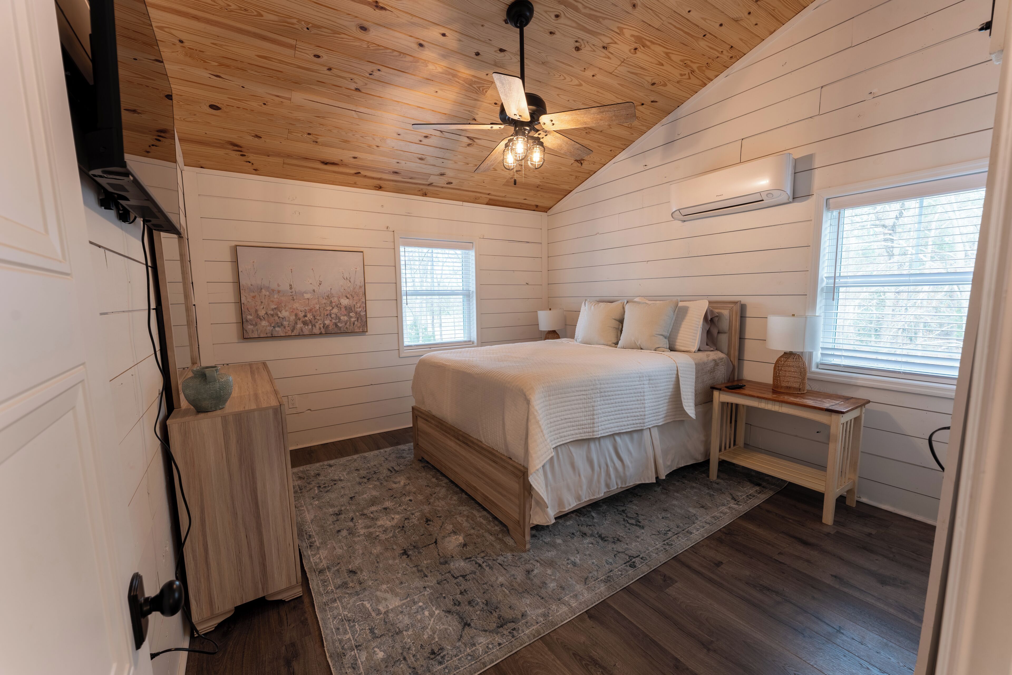A bedroom with a wooden ceiling, a bed with white bedding, a bedside table, a nightstand, a painting on the wall, and a window with blinds.