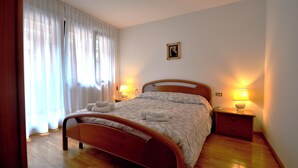 Un letto rifatto con lenzuola e asciugamani bianchi, un comodino con una lampada e una finestra con tende leggere.