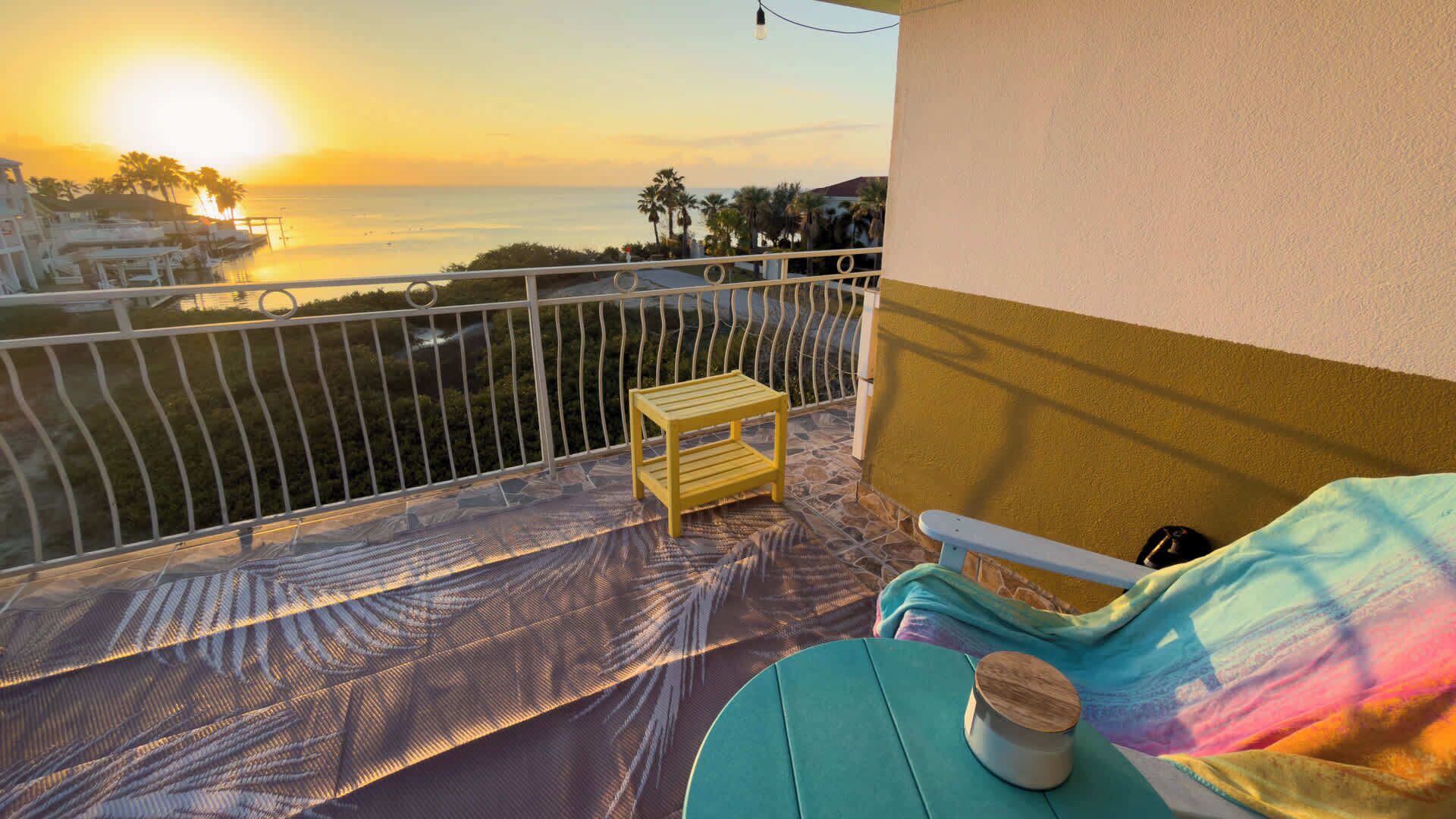 A balcony with a view of the sunset over the sea, a small wooden table, and a colorful sofa.