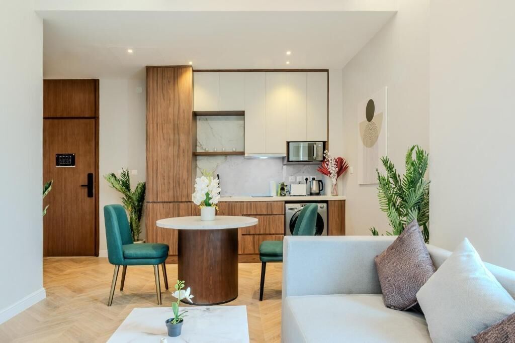 A modern kitchen with wooden cabinets, a dining area with a round table and chairs, and a living room with a sofa and cushions.