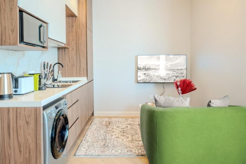 A compact kitchen with a washing machine, a microwave, and a sink. There is a green sofa and a framed picture on the wall.