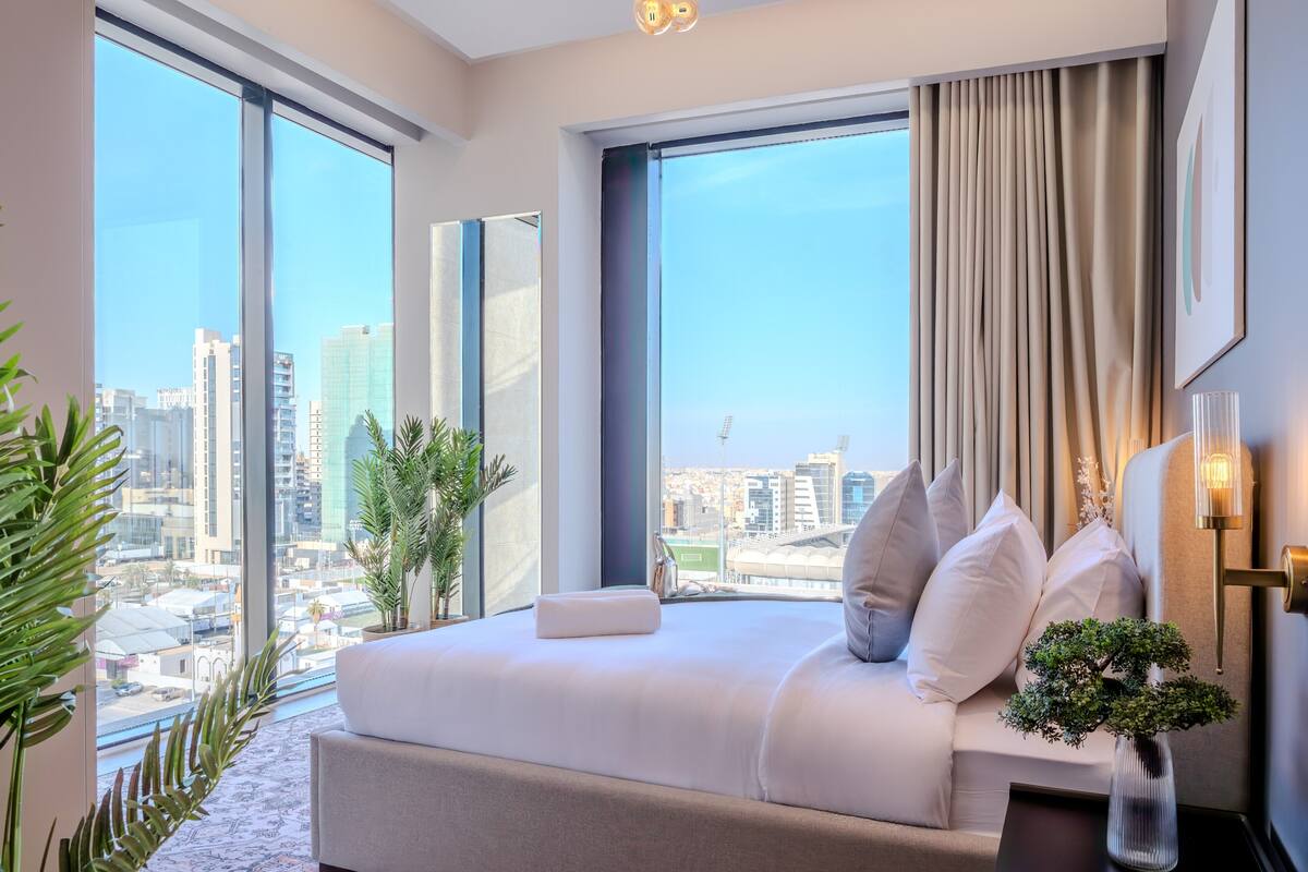 Quarto de hotel moderno com uma cama grande, vista para a cidade e uma planta em vaso.