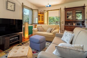 A living room with a sofa, ottoman, and television, a cabinet with glass doors, and a clock on the wall. - Villa Angiolina - Ideal for 5 people with A/C and Wi-Fi (Sardegna)