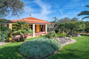 A house with a red-tiled roof, a covered porch, and a garden with various plants and a lawn. - Villa Angiolina - Ideal for 5 people with A/C and Wi-Fi (Sardegna)