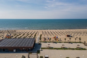 Uma praia com fileiras de guarda-sóis e palmeiras, um edifício com painéis solares e vista clara para o oceano.