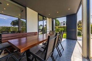 Uma área moderna para refeições ao ar livre com mesa e cadeiras de madeira, amplas janelas e vista para o verde ao redor.