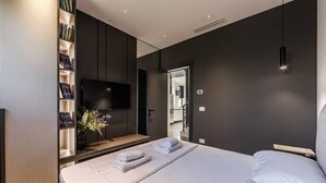 A modern bedroom with a large bed, a bookshelf, a flat-screen TV, and a view of the hallway.