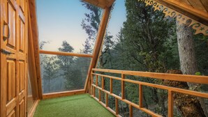 A room with a wooden balcony, a glass wall, and a view of a forest.