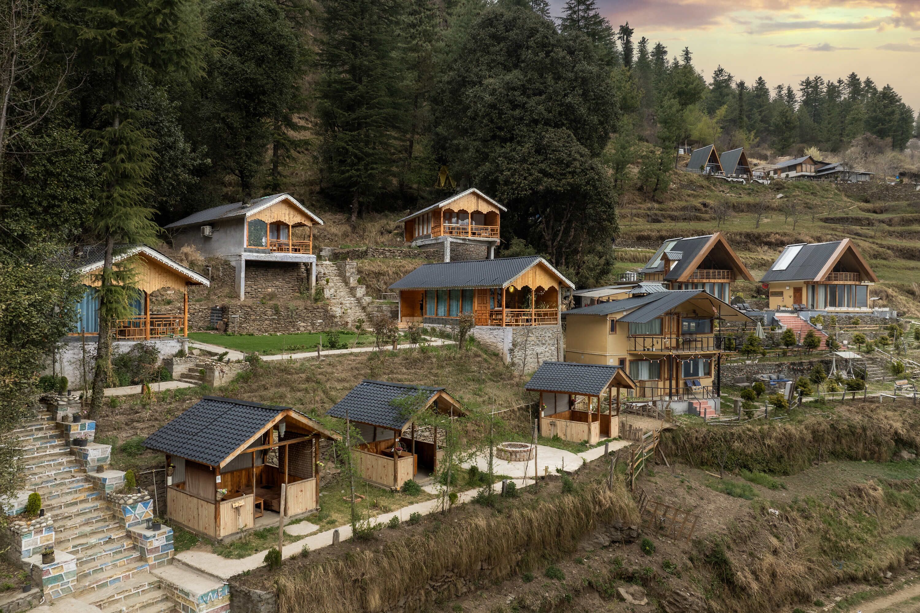 A cluster of wooden cabins with stone foundations, set on a hillside with a forest backdrop.