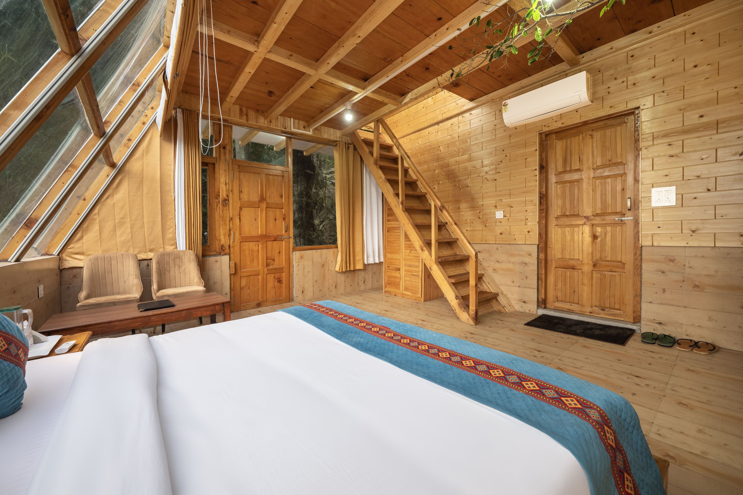 A wooden cabin with a bed, a desk, a chair, a staircase, and a large window.