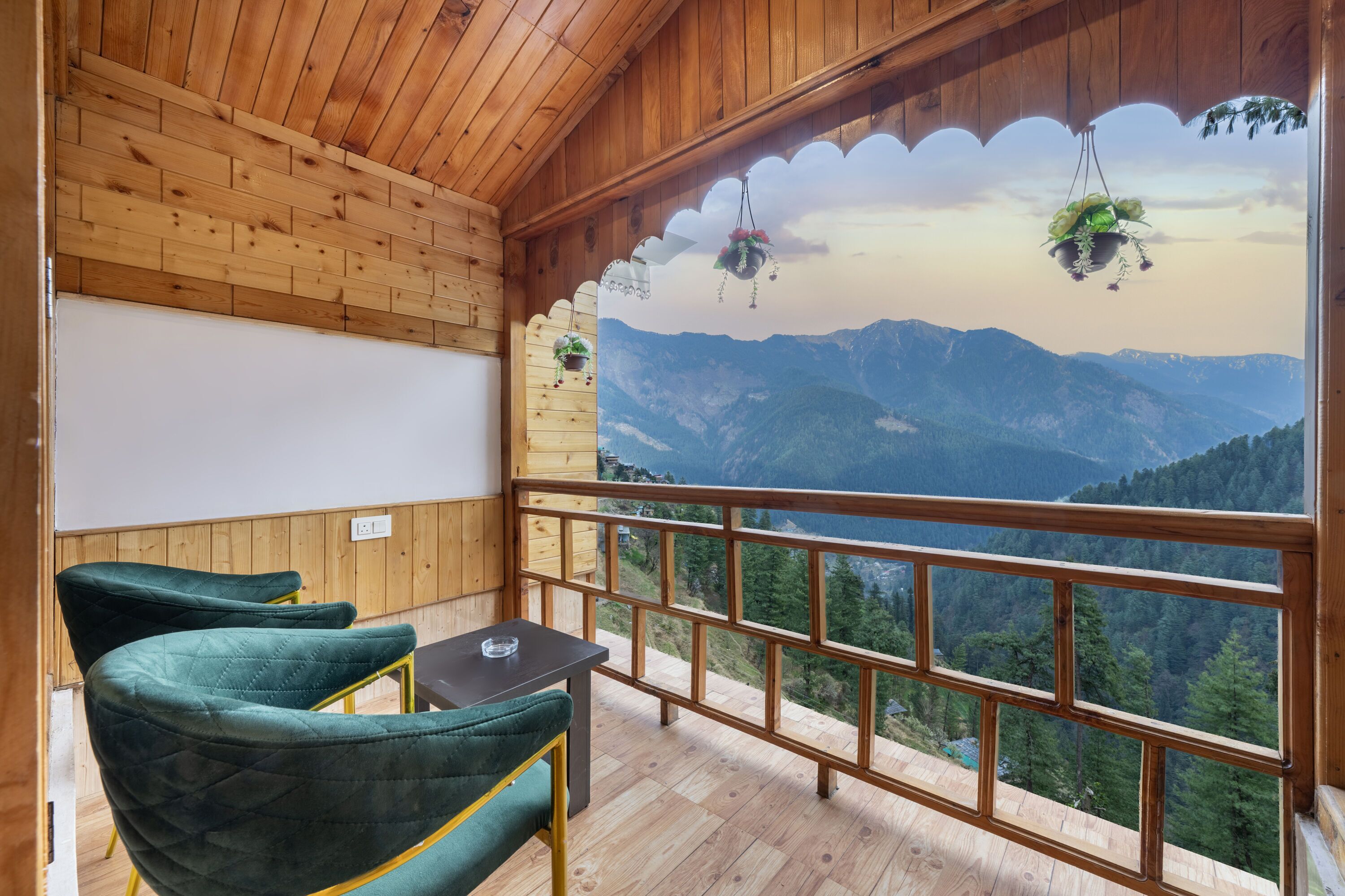 A wooden balcony with a view of mountains, two green upholstered chairs, and hanging plants.