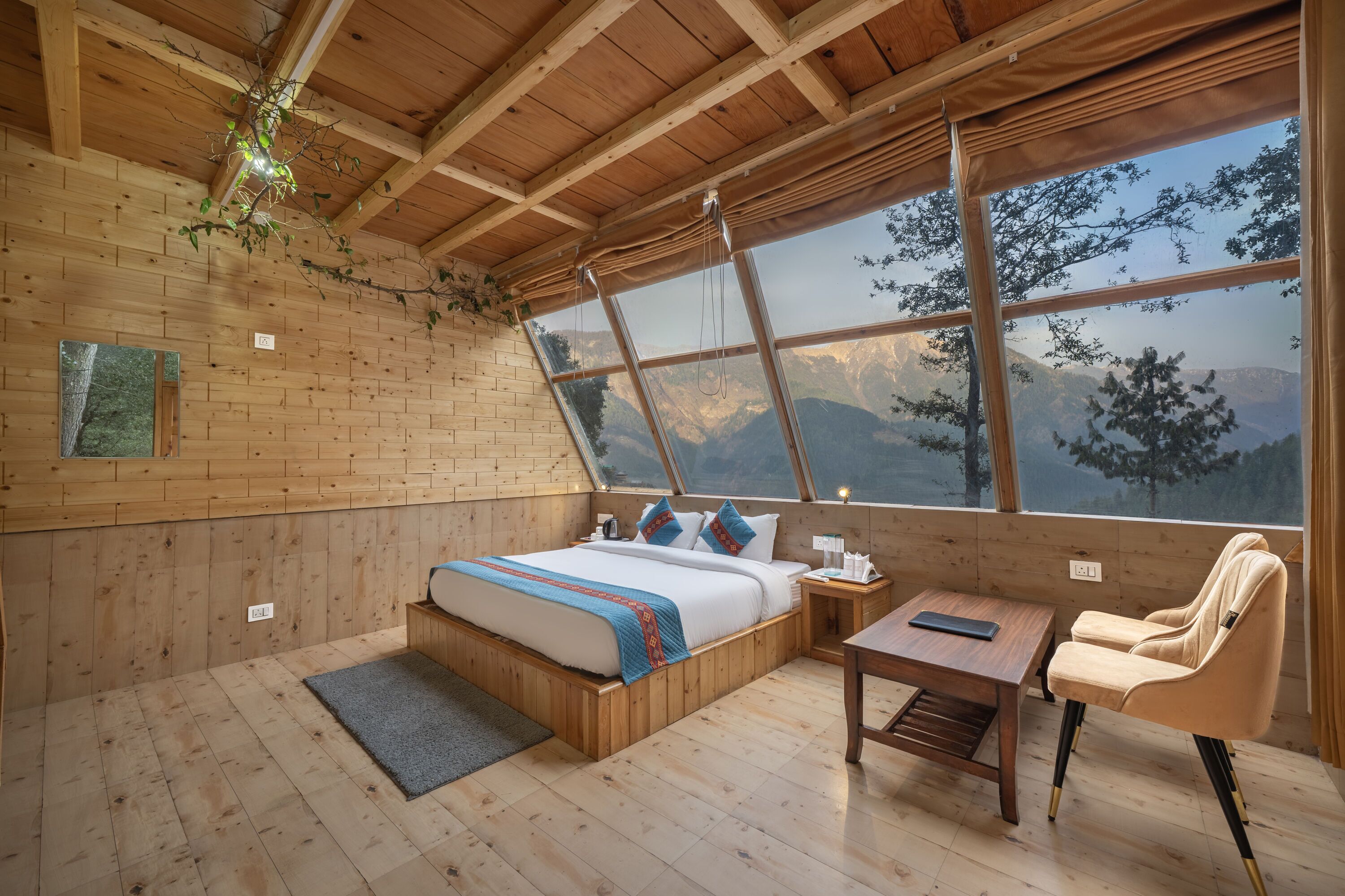 A wooden cabin bedroom with a bed, bedside table, chair, and a large window overlooking mountains.