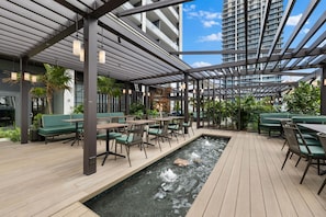 Eine Dachterrasse mit Außenbestuhlung, einem kleinen Wasserspiel und Blick auf ein hohes Gebäude.