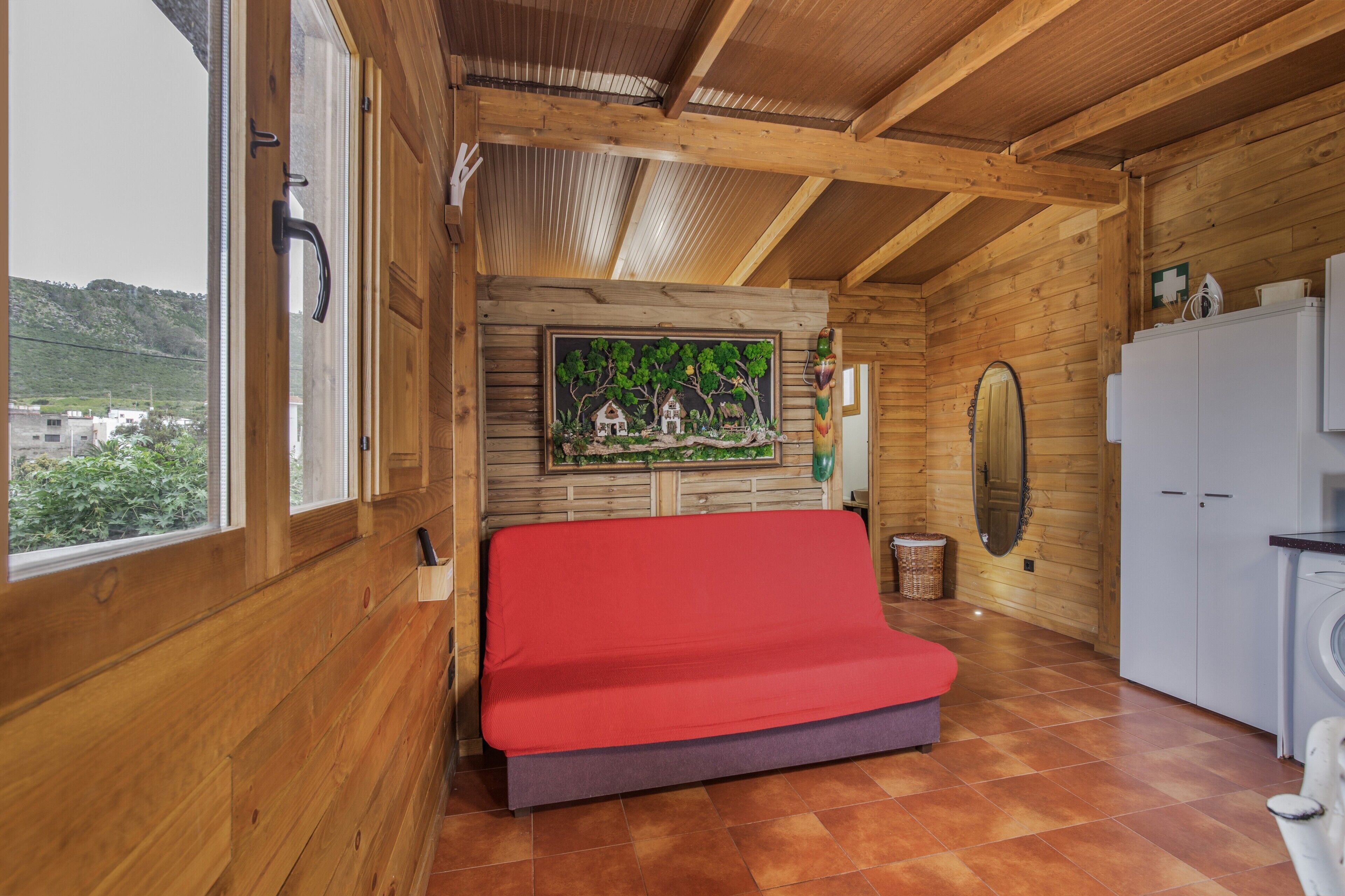 Un interior de madera con una cama, una lavadora y vistas por una ventana.