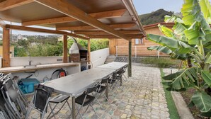 Ein überdachter Außenbereich mit einer Steinterrasse, einem Grill und einem Tisch mit Stühlen.