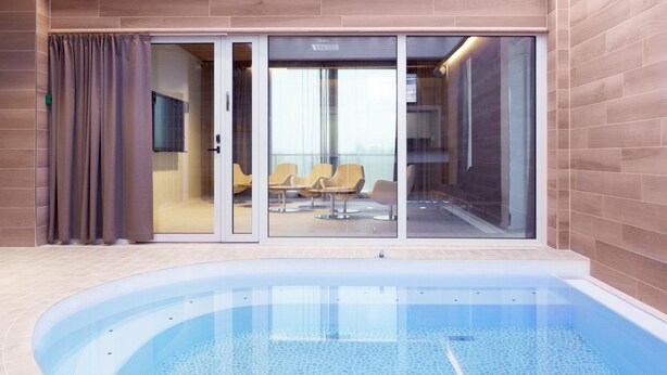 Een modern binnenzwembad met helderblauw water, een glazen deur naar een zithoek met stoelen, en een betegelde vloer.