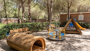 A wooden play structure, a wooden bench, and a small table with chairs in a sandy area surrounded by trees and wooden cabins. - Mobilhome im Ferienpark mit Grosser Poolanlage (Castiglione della Pescaia)
