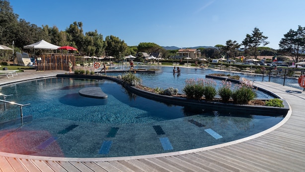 A large, circular infinity pool with a wooden deck surrounding it, featuring lounge chairs and umbrellas. - Mobilhome im Ferienpark mit Grosser Poolanlage (Castiglione della Pescaia)