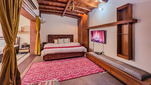 Un dormitorio acogedor con techo de madera, una cama grande con colcha estampada, un televisor montado en la pared y un banco con asiento de mimbre.
