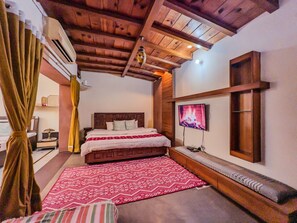 A cozy bedroom with a wooden ceiling, a large bed with a patterned bedspread, a TV mounted on the wall, and a bench with a woven seat. - The Blush house 3 BHK (Jaipur)