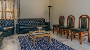 Una sala de estar con un sofá verde oscuro, dos sillas de madera, una mesa de centro de vidrio y alfombras estampadas.