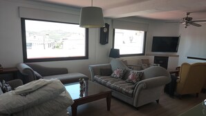 En stue med sofa, lænestole, sofabord og fjernsyn. Store vinduer giver udsigt til bygninger.