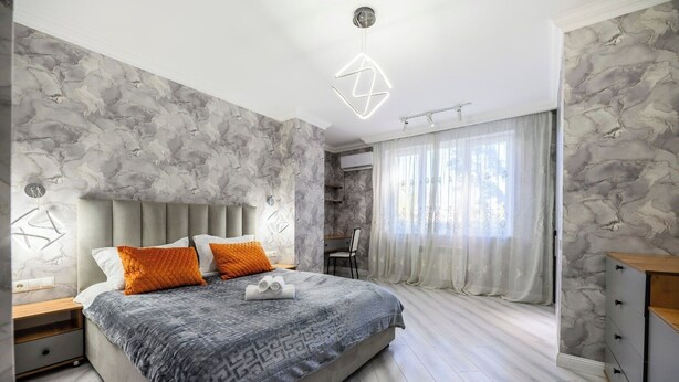 A modern bedroom with a marble-patterned wall, a bed with a grey and orange quilted bedspread, a wooden nightstand, and a desk with a chair.