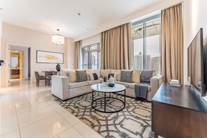 A modern living room with a sofa, coffee table, and TV. There is a dining area with chairs and a table. Large windows offer a city view. - Stylish 2BR with Burj Khalifa Views, Minutes from Marine Transport Station (Dubai)
