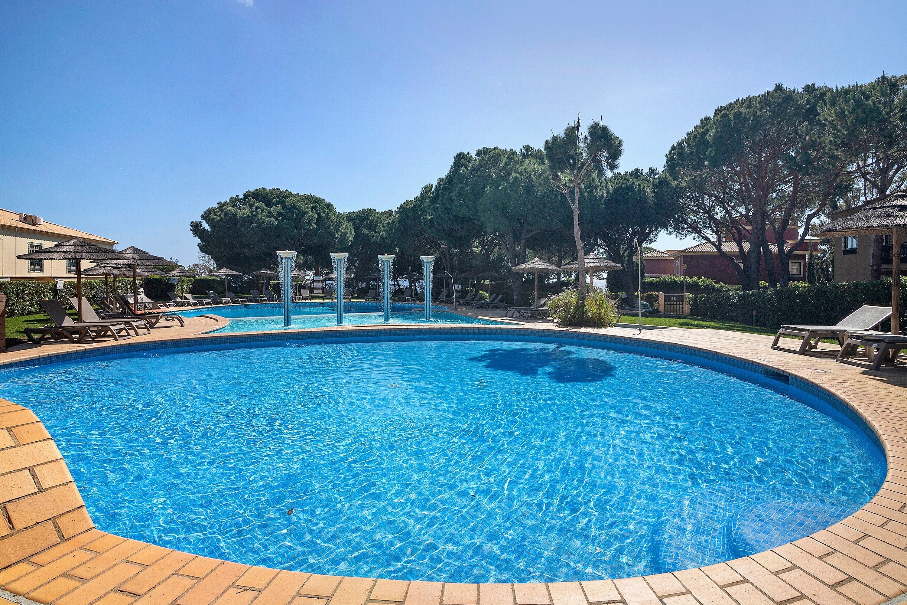 A swimming pool with lounge chairs and umbrellas, surrounded by trees and buildings.