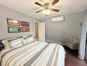 Ein Schlafzimmer mit einem Bett, Deckenventilator, Wandklimaanlage, gerahmten Bildern und einer Tür.