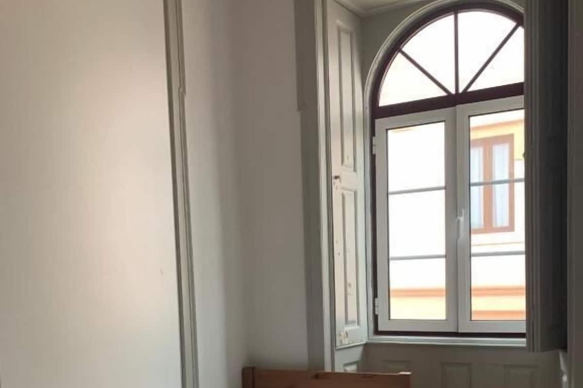 Una cama individual con cabecera de madera, un taburete de madera pequeño y una ventana con vistas a un edificio.