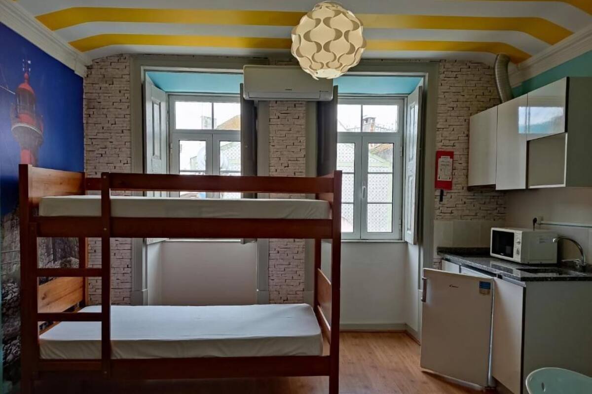 Una cama litera en una habitación con pared de ladrillo, una ventana, un área de cocina con microondas y una lámpara colgante.