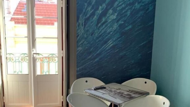 A room with a wooden floor, a white table with chairs, a large window with a view of buildings, and a blue textured wall. - Meeting Hostel (Figueira da Foz)