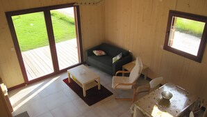 A room with a wooden floor, a small white table, a green sofa, a white chair, and a window with a view of a grassy area. - Furnished Cottage in the Land of Castles – Ideal for Cycling (Mont-près-Chambord)