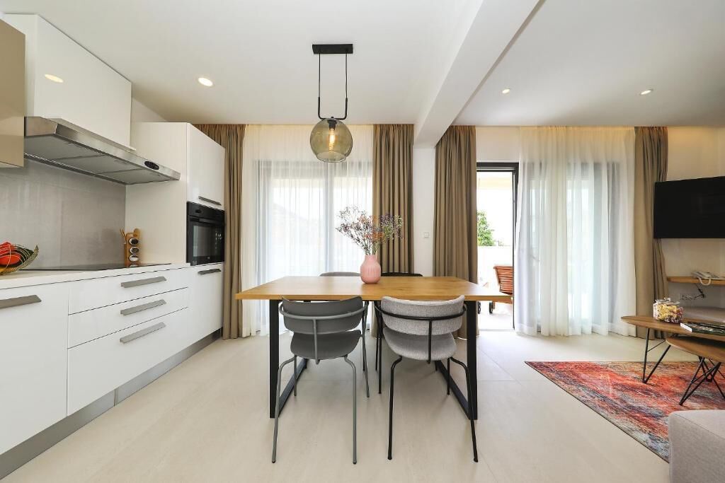 A modern kitchen with white cabinets, a wooden dining table, two chairs, a hanging light fixture, and a TV on the wall.
