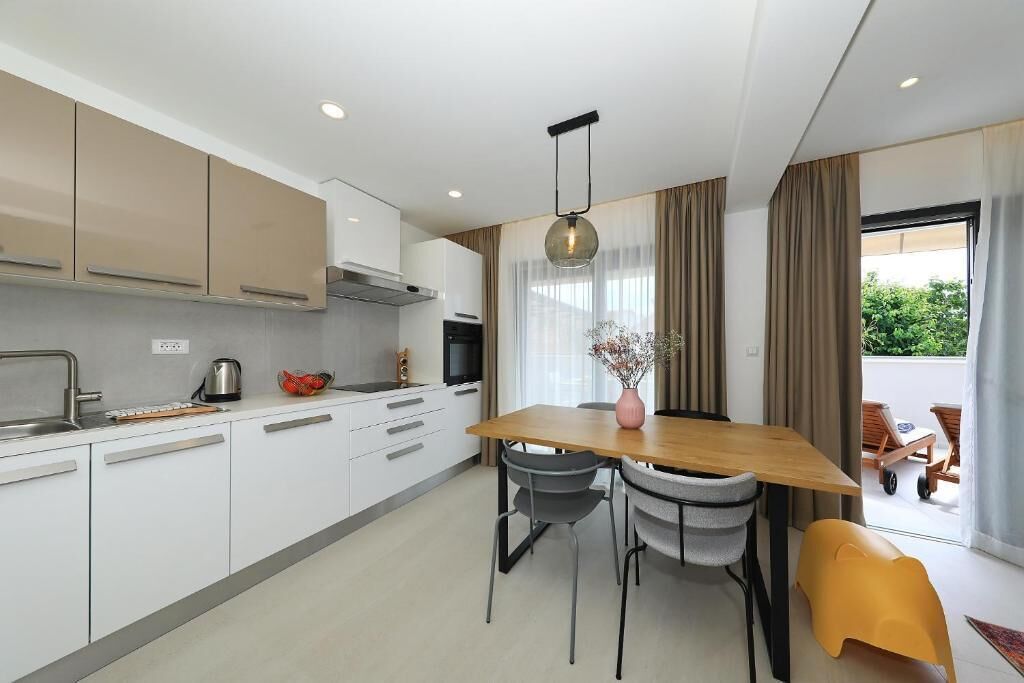 A modern kitchen with white cabinets, a wooden dining table, and a view of the outdoors.