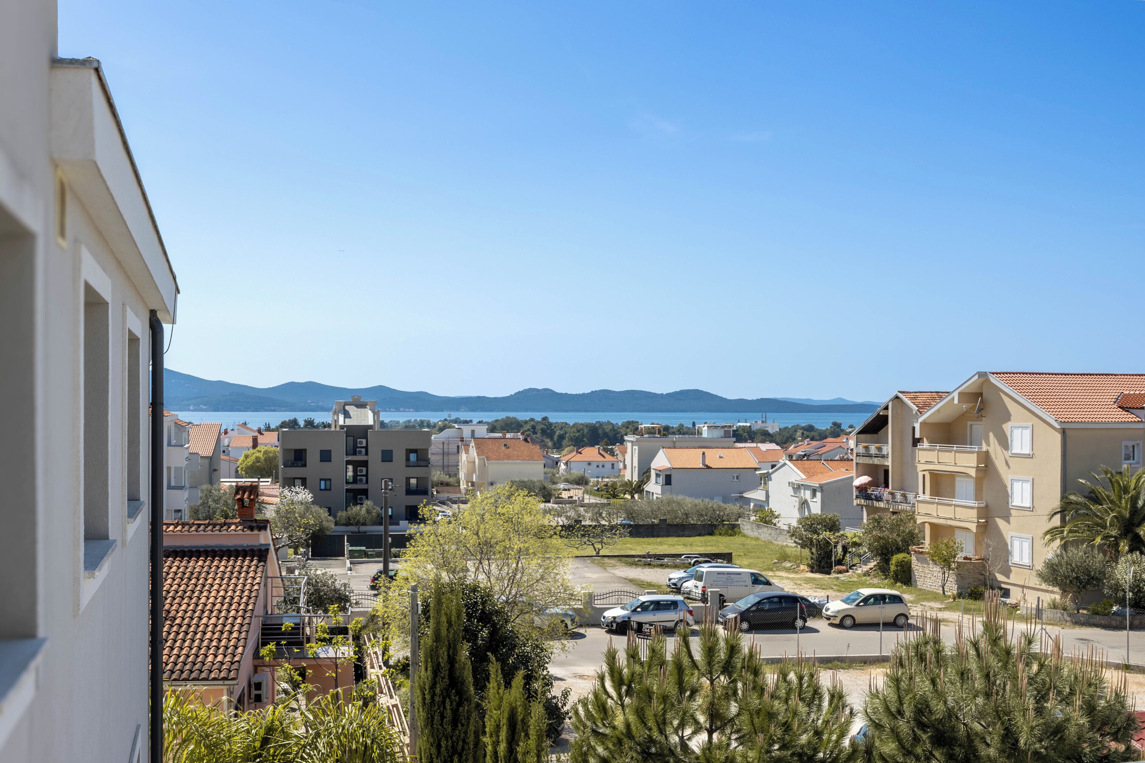 A residential area with buildings, parked cars, and a clear view of the sea and mountains in the distance.