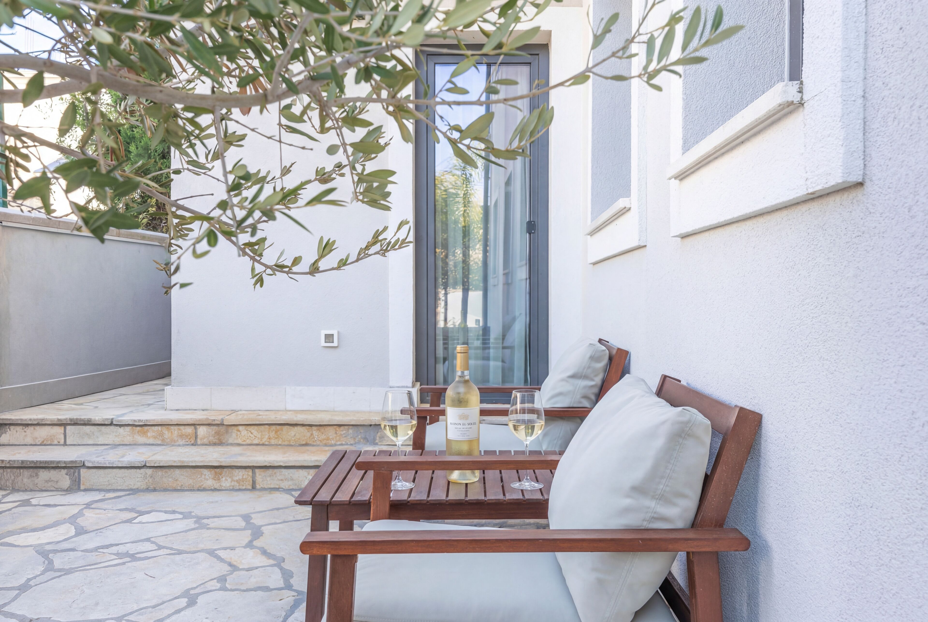 A patio with a wooden table and two chairs, a bottle of wine, and two glasses.