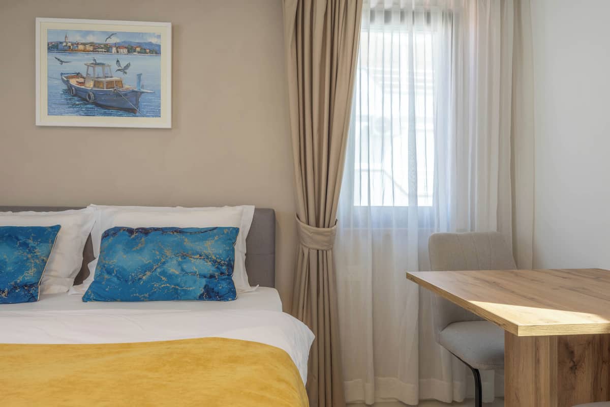 Ein Hotelzimmer mit einem Bett, zwei blauen Kissen, einem Holztisch, einem Stuhl und einem Bild eines Bootes an der Wand.