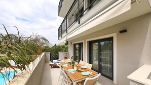 Un balcón con una mesa de comedor dispuesta para cuatro personas, rodeado de sillas blancas, una piscina y un edificio con balcón.