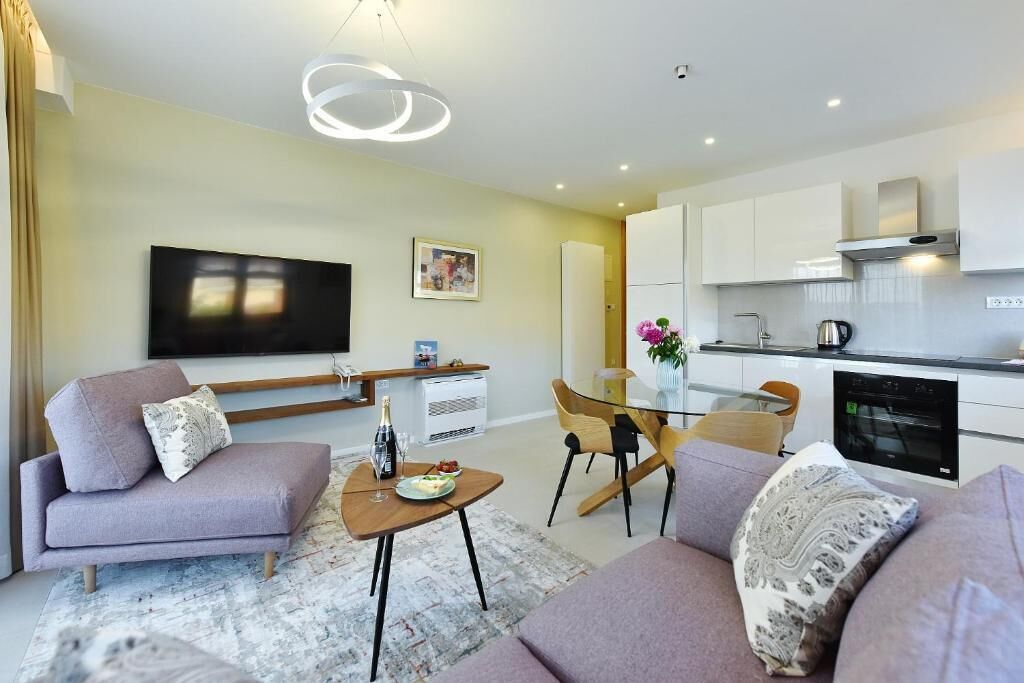 Un salon moderne avec un canapé, une table basse et un téléviseur. On y trouve également un coin repas avec une table et des chaises en verre. La cuisine est visible et se distingue par ses meubles blancs et sa cuisinière.