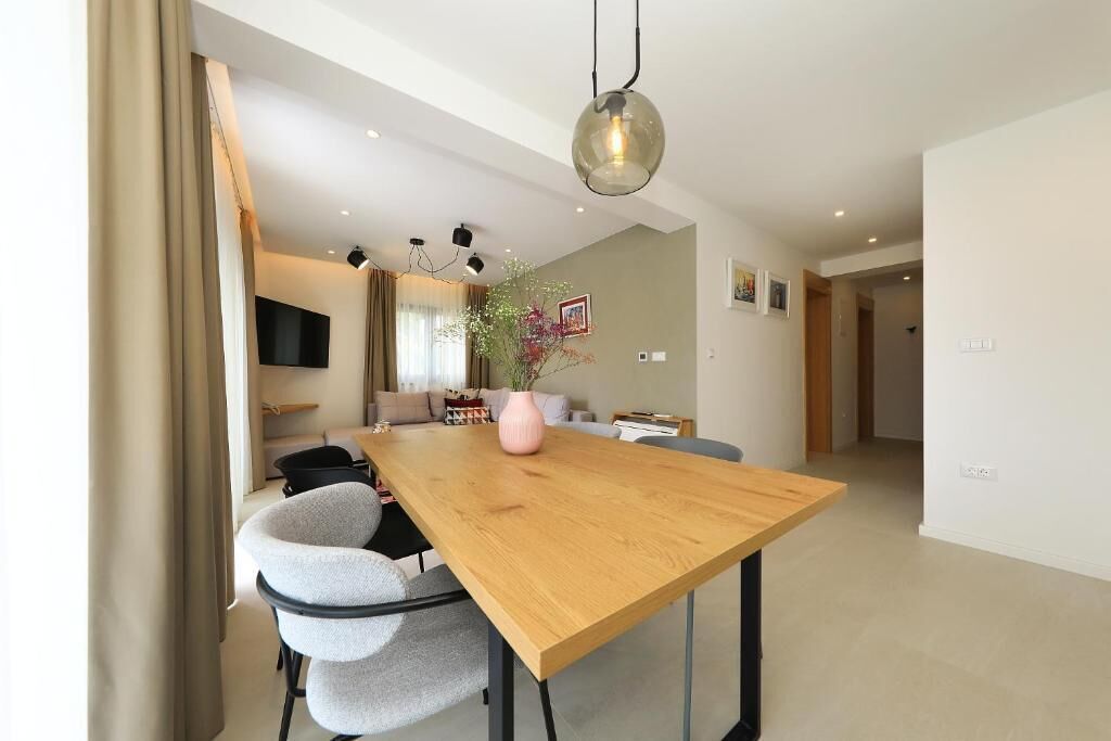 Ein moderner Essbereich mit Holztisch, grau gepolsterten Stühlen, einer rosafarbenen Vase und einer hängenden Leuchte.