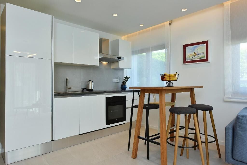 A modern kitchen with white cabinets, a built-in oven, and a dining area with a wooden table and stools.