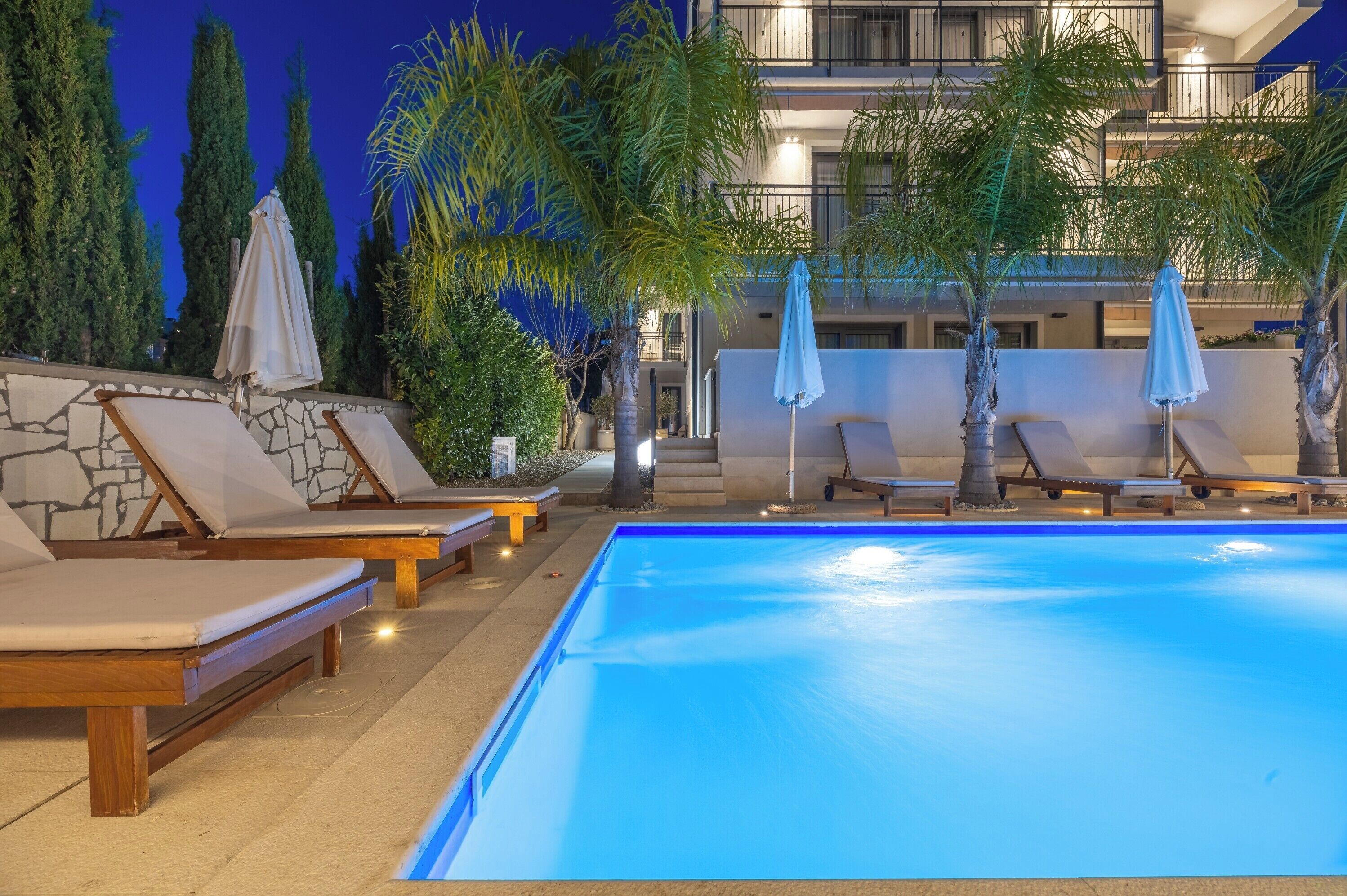Una piscina con sillones de descanso y sombrillas, rodeada de árboles y un edificio con balcones.