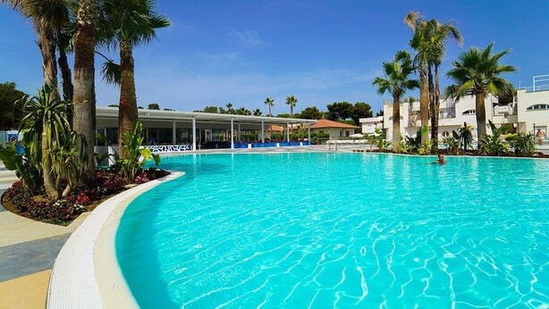 A large swimming pool with palm trees, a covered area, and buildings in the background. - FRUIT VILLAGE PAESTUM Coral Beach (Capaccio-Paestum)
