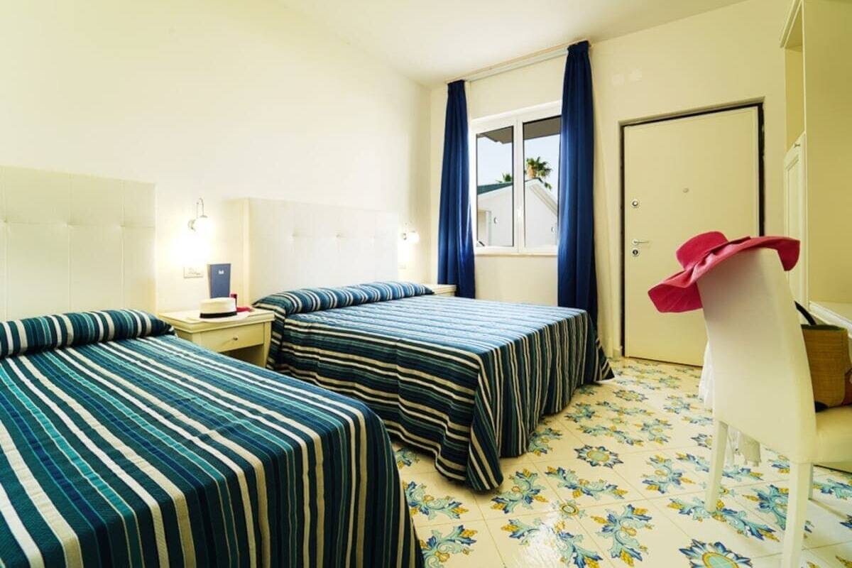 A hotel room with two beds, a desk, a chair, a window with curtains, and a door.