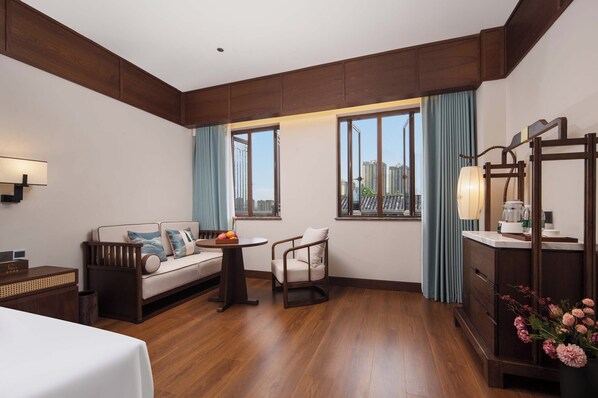 A hotel room with a wooden floor, a sofa, a chair, a small table with fruit, a lamp, a dresser, and a view of the city through large windows. - Chongqing Menghua Lushe Hotel (Chongqing)