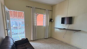Ein Wohnzimmer mit einem dunkelbraunen Sofa, einem Deckenventilator, einem an der Wand montierten Fernseher und einem Fenster mit Vorhängen.
