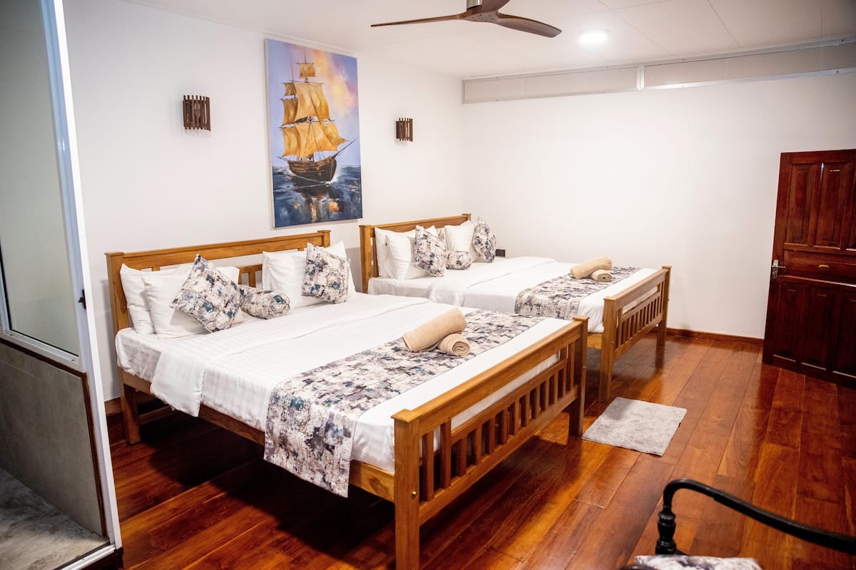 Una habitación de hotel con dos camas de madera, un banco de madera, un ventilador de techo y un cuadro de un barco en la pared.